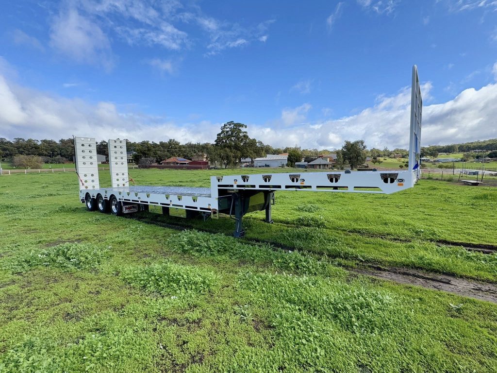 Side view of FreightWest next generation heavy duty triaxle drop deck trailer parked on grass with trees in the background
