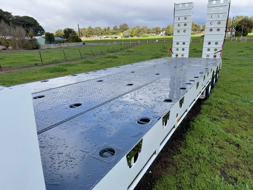 Looking down the length of the FreightWest drop deck trailer from the rear on a dirt yard with vehicles in the background