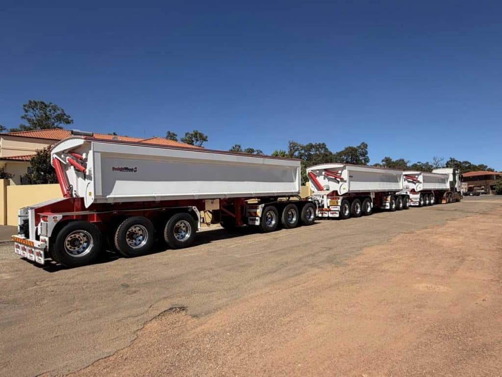 A line of FreightWest triple side tipper trailers parked in a yard under a clear blue sky