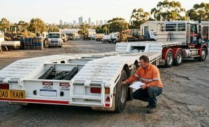A man in a high-vis shirt crouches to inspect the wheels of a large drop deck trailer in a Perth industrial yard.