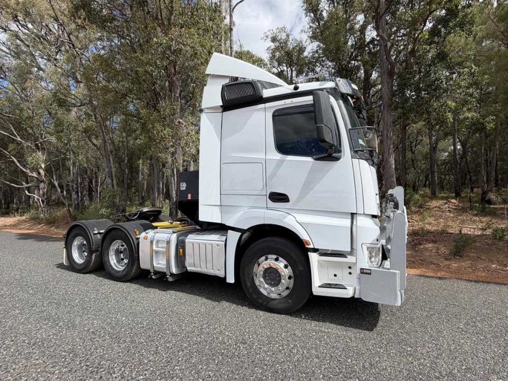Side profile view of the white semi-truck prime mover parked outdoors next to trees